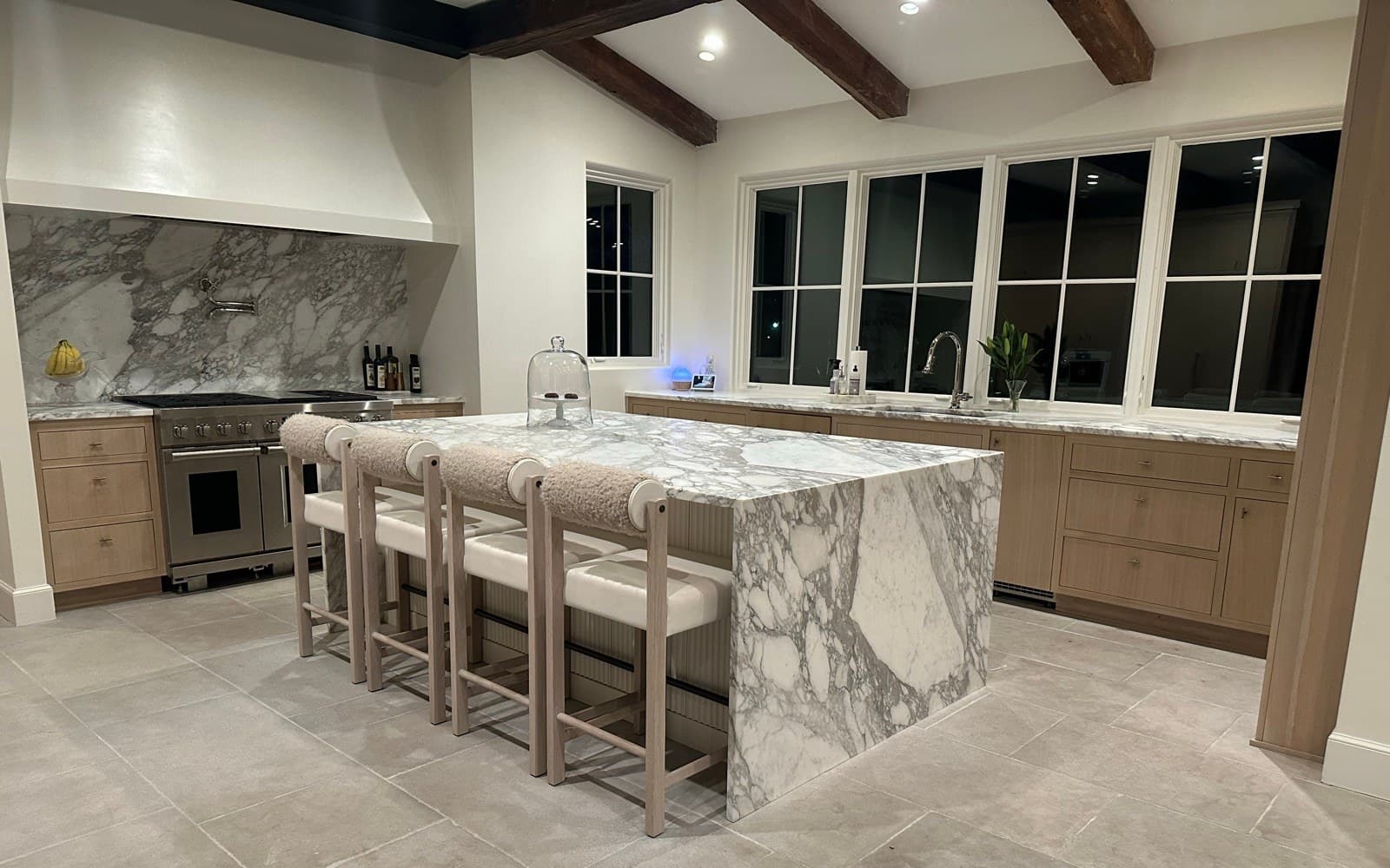 A marble kitchen island with four chairs, oak cabinetry, and exposed wood beams